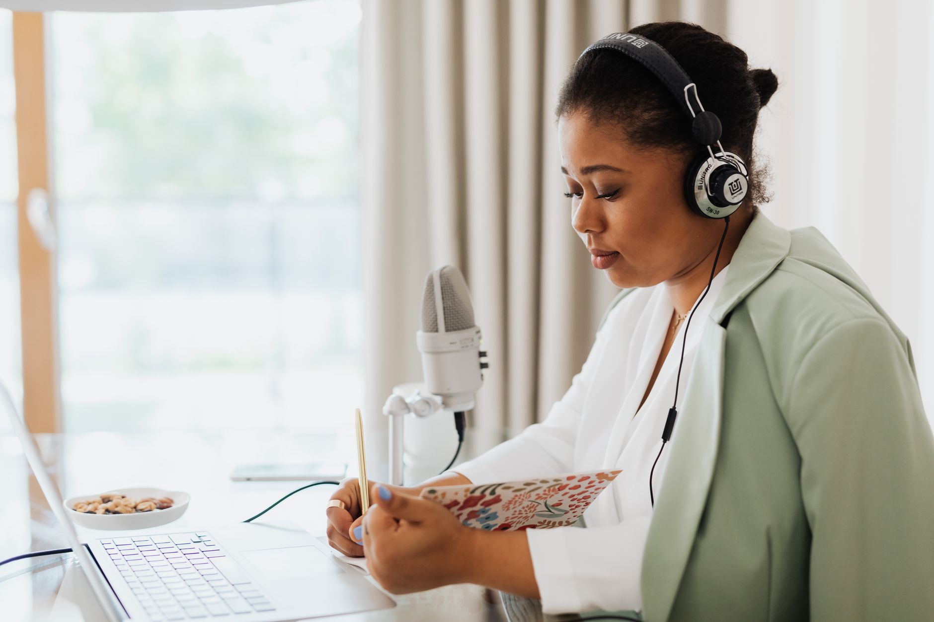 Una donna con le cuffie prende appunti su un quaderno davanti a un portatile e a un microfono da studio